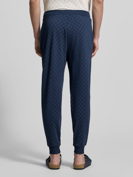 Męski model nosi Sweatpants mit Logo-Muster und elastischem Bund von JOOP! Collection z bawełny, krój regular fit, wzór z logo, długość wewnętrzna nogawki w rozmiarze M: 75 cm, widok całej sylwetki od tyłu