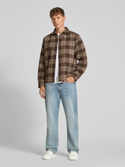 Männermodel trägt Freizeithemd mit durchgehender Knopfleiste von Selected Homme, kariertes Allover-Muster in Braun, Knöpfe, Kragen, Langarm, dazu Jeans und Sneaker, Ganzkörperansicht von vorne.