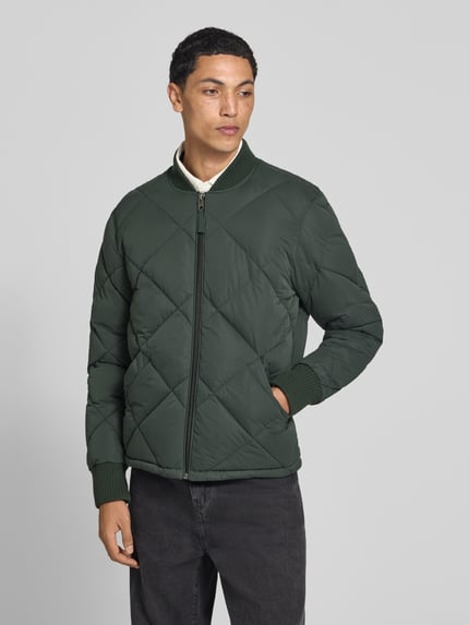 Männermodel trägt Regular Fit Steppjacke mit Stehkragen van Superdry in groene Stiksels look, steekzakken, ritssluiting, elastische mouwboordjes, ruglengte maat M 69 cm, schouderbreedte 46 cm, borstwijdte 58 cm, mouwlengte 59 cm. Ganzkörperansicht van voren.