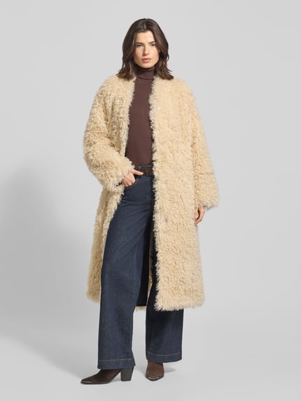 Frauenmodel trägt Mantel aus Kunstfell Modell 'Carolina' von STAND STUDIO in Beige, lockerer Schnitt, V-Ausschnitt, Ganzkörperansicht von vorne.