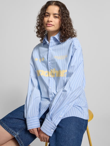 Frauenmodel trägt Relaxed Fit Bluse mit Label-Print von ANOTHER COTTON LAB aus reiner Baumwolle, blau mit weißen Streifen, Hemdblusenkragen, Knopfleiste, Vorderansicht, Nahaufnahme Oberkörper.