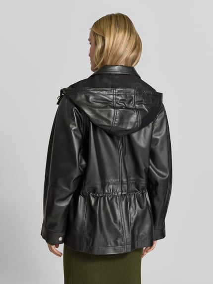 Frauenmodel trägt Funktionsjacke Modell 'GROOVE' von IRO in schwarzer, glänzender Lederoptik mit Kapuze, Struktur und metallischen Akzenten, Ganzkörperansicht Rückansicht.