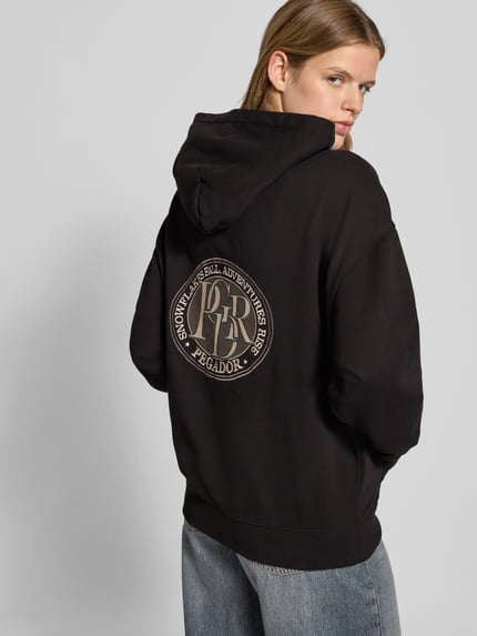 Vrouwenmodel draagt oversized zwarte hoodie met kangoeroezak, logo op de rug, Oversized Hoodie met Känguru-Tasche Modell 'AVERSA' van Pegador, katoenmix, Ganzkörperansicht Rückansicht.