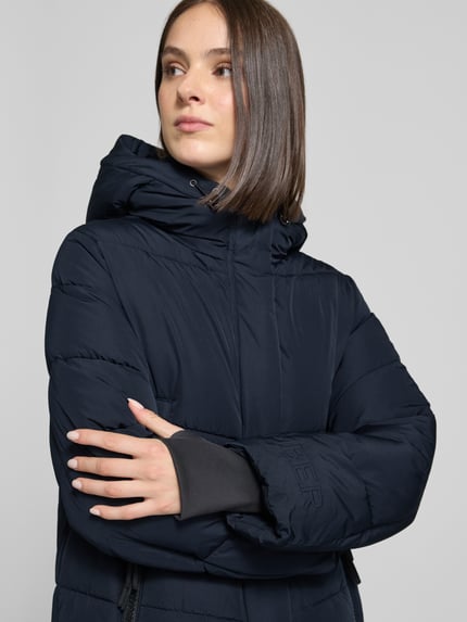 Frauenmodel trägt Steppmantel mit Reißverschlusstaschen von Amber & June in Dunkelblau, wattierte Oberfläche, Kapuze mit Tunnelzug, Nahaufnahme Oberkörper, Regular Fit, verdeckter Reißverschluss, seitliche Reißverschlusstaschen, Damenmodell Größe 36, KI-modifiziertes Bild.