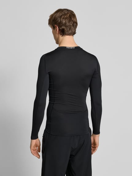 Męski model nosi Longsleeve mit Label-Print od adidas Training, kolor czarny, materiał z domieszką stretchu, krój slim fit, nadruk z logo z tyłu, długość pleców w rozmiarze M: 65 cm, widok całej sylwetki z tyłu