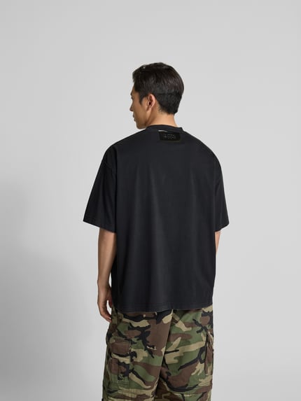 Mężczyznamodel nosi męski t-shirt z czarnej bawełny od VETEMENTS, krój relaxed fit, okrągły dekolt, detal z logo, z krótkim rękawem, długość tyłu w rozmiarze M: 77 cm, widok z tyłu.
