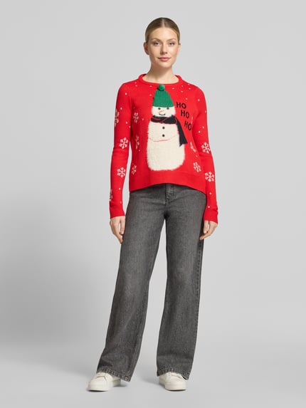 Frauenmodel trägt Pullover mit flauschigem Schneemann-Motiv von VERO MODA in Rot mit Schneeflocken-Print, Strickoptik, Ganzkörperansicht von vorne.