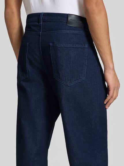 Männermodel trägt Wide Fit Jeans aus reiner Baumwolle von MAISON KITSUNÉ in dunkelblauer Waschung, sichtbare Gesäßtaschen, Ganzkörperansicht Rückansicht. Kontext: Herren Jeans, Wide Fit, Knopf- und Reißverschluss, 5-Pocket-Design, gleichmäßige Waschung, Innenbeinlänge 78 cm, Bundweite 45 cm.