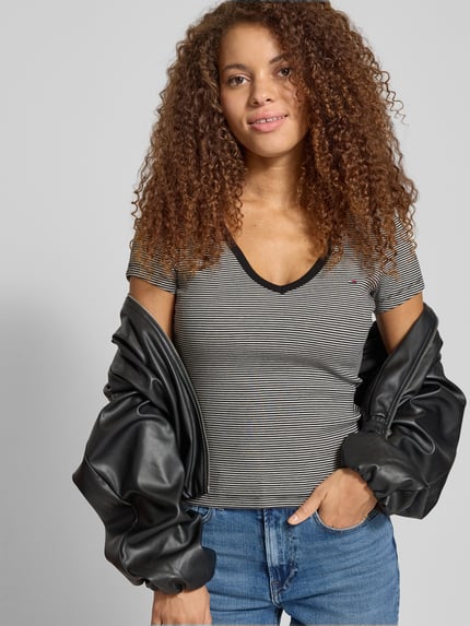 Modelka kobieca nosi Slim Fit T-Shirt aus Baumwoll-Mix von Tommy Jeans, cienki prążkowany materiał, logo na piersi, V-dekolt, krótki rękaw, jednolity kolor, widok całej sylwetki z przodu.