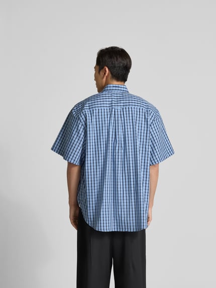 Männermodel trägt Oversized Freizeithemd mit Brusttasche von VETEMENTS in Blau mit Karomuster, Reine Baumwolle, Oversized-Fit, Kurzärmelig, Kentkragen, Rückenlänge bei Größe M 82 cm, Schulterbreite 55 cm, Ärmellänge 29 cm, Ganzkörperansicht Rückansicht.