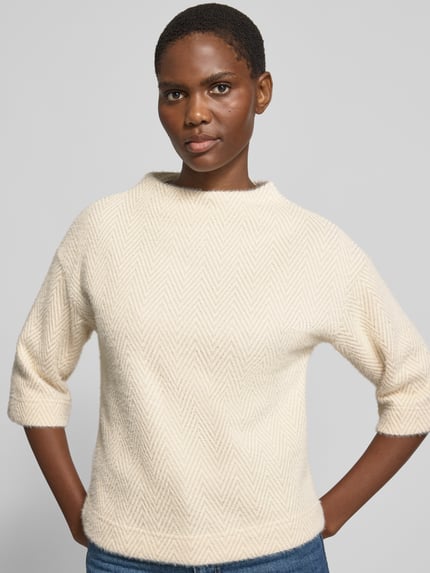 Modelka kobieca ma na sobie sweter Pullover mit Stehkragen und 3/4-Arm od zero, Mieszanka materiałów z dodatkiem streczu, krój regular fit, stójka, rękaw o dł. 3/4, jasny kolor, tekstura w jodełkę, zbliżenie na górną część ciała, widok z przodu.