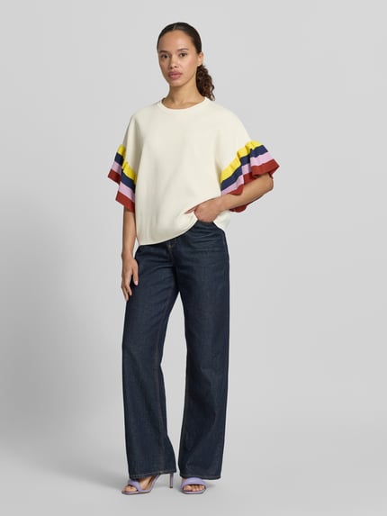 Frauenmodel trägt Boxy Fit Pullover aus Viskose-Mix Modell 'JANUARY' von Essentiel Antwerp in Creme mit bunten Streifen an den Ärmeln, weite Passform, glatte Textur, kombiniert mit weiter Jeans, Ganzkörperansicht von vorne.