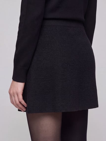 Frauenmodel trägt schwarzen Mini-Rock mit Taillenpasse von PIECES, A-Linie, strukturierte Strickoptik, Ganzkörperansicht Rückansicht.