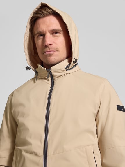 Männermodel trägt Regular Fit Jacke mit Stehkragen von bugatti in Beige mit Logo-Detail, Zweiwege-Reißverschluss, Kapuze, Nahaufnahme Oberkörper.