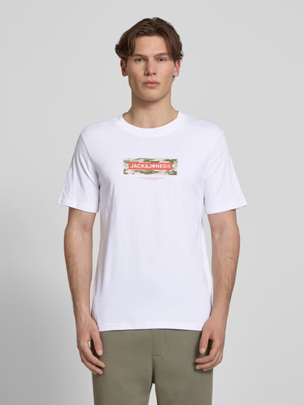 Męski model nosi T-Shirt mit Logo und Rundhalsausschnitt od Jack & Jones, czysta bawełna, krój regular fit, długość tyłu w rozmiarze M: 68 cm, biała koszulka z nadrukiem, widok całej sylwetki z przodu.