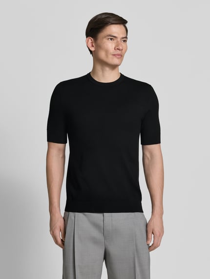 Männermodel trägt Slim Fit T-Shirt aus Strick von HUGO, schwarze Strick-Optik, Rundhals, Kurzarm, Label-Stitching-Applikation, Ganzkörperansicht von vorne.