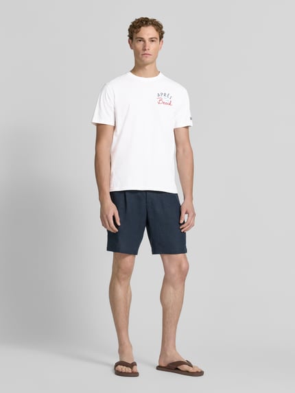Männermodel trägt Herren T-Shirt mit Label-Prints Modell 'PORTLAND' von MC2 Saint Barth aus reiner Baumwolle, Regular-Fit, Rundhalsausschnitt, Vorder- und Rückseite mit Label-Print, Ärmel mit Label-Stitching, Farbe Weiß, kombiniert mit dunkelblauer Shorts, Ganzkörperansicht von vorne.