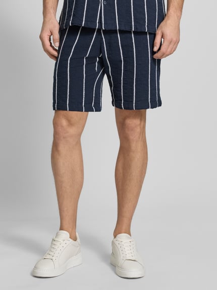 Mężczyzna nosi Shorts mit elastischem Bund und Tunnelzug von MCNEAL, bawełna, krój regular fit, szerokość w rozmiarze M 42 cm, długość nogawki 20 cm, ciemnogranatowe z białymi paskami, faktura lekko marszczona, widok z przodu na całe ciało.