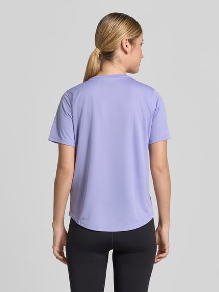 Frauenmodel trägt T-Shirt mit Rundhalsausschnitt und Logo-Print von ASICS in zartem Lila, gerader Schnitt, leicht abgerundeter Saum, Rückenlänge 60 cm, Ganzkörperansicht Rückansicht.