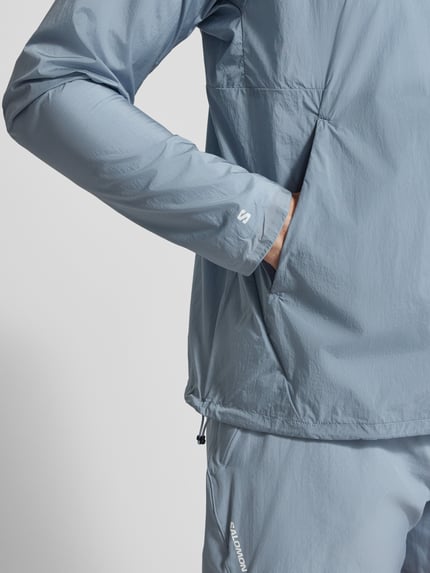 Męski model nosi Jacke mit Logo und Eingrifftaschen od Salomon, jasnoniebieski, logo, zamek błyskawiczny, wpuszczane kieszenie, krój regular fit, stójka, długość tyłu w rozmiarze M: 70 cm, szerokość ramion w rozmiarze M: 43 cm, szerokość klatki piersiowej w rozmiarze M: 35 cm, długość rękawów w rozmiarze M: 67 cm, detal kurtki, zbliżenie na górną część ciała
