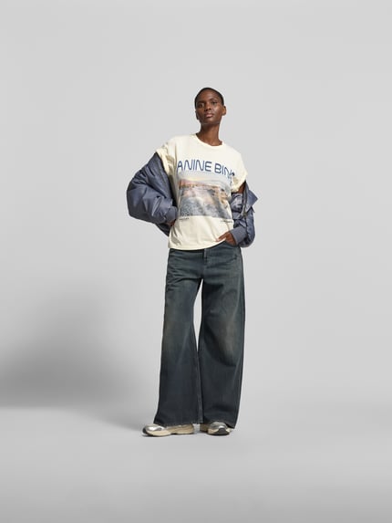 Frauenmodel trägt Regular Fit T-Shirt mit Logo-Print von Anine Bing aus reiner Baumwolle, heller Stoff mit Motiv-Print, Rundhalsausschnitt, Rückenlänge 60 cm bei Größe S, Ganzkörperansicht von vorne.