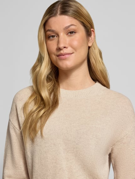 Kobiece model prezentuje Strickpullover mit überschnittenen Schultern od Christian Berg Woman, jasny beż, długie rękawy, luźny fason, prążkowane wykończenia, okrągły dekolt, długość tyłu 60 cm, długość rękawa 58 cm, zbliżenie na górną część ciała.