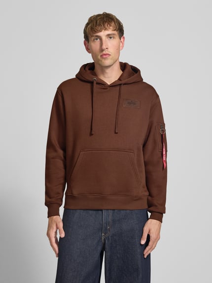 Männermodel trägt Hoodie mit Känguru-Tasche von Alpha Industries in Braun, Baumwoll-Mix, lockerer Schnitt, Ganzkörperansicht von vorne.