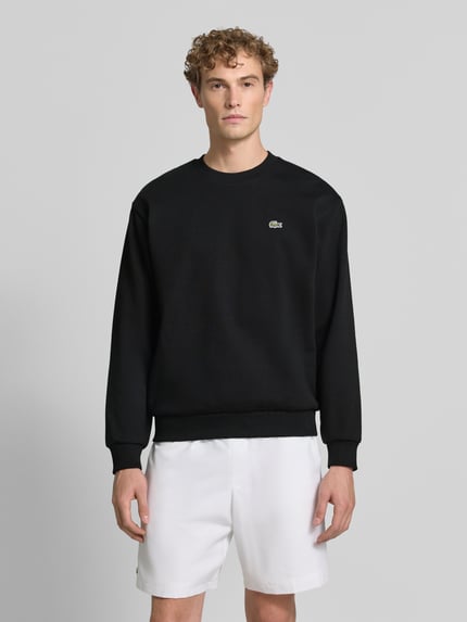 Männermodel trägt schwarzes Regular Fit Sweatshirt aus Baumwoll-Mix von Lacoste, glatte Struktur, weiße Shorts, Ganzkörperansicht von vorne. Herren Sweatshirt von Lacoste