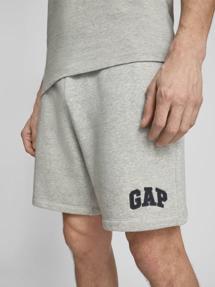 Mężczyzna nosi Sweatshorts mit elastischem Bund von GAP z mieszanki bawełny, krój relaxed fit, logo GAP, elastyczny pas, szerokość w rozmiarze M: 41 cm, długość wewnętrzna nogawki w rozmiarze M: 20 cm, jasnoszare, gładka tekstura, z przodu detal logo, zbliżenie na dolną część ciała, perspektywa z przodu.