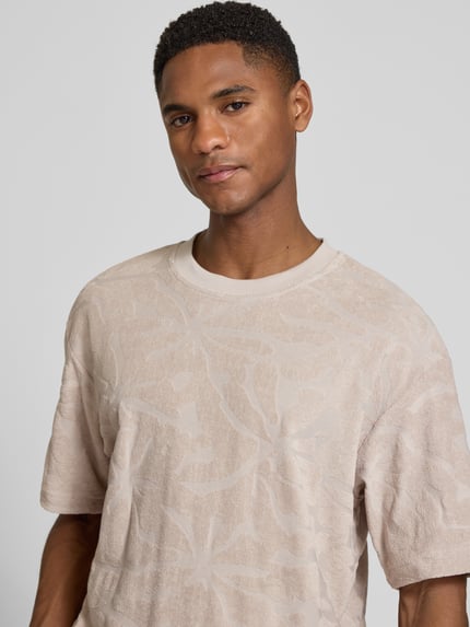 Männermodel trägt Regular Fit T-Shirt aus Baumwoll-Mix Modell 'TE_FLOWER' von BOSS Orange mit Optik: Allover-Muster, Struktur, Label-Applikation, Rundhals, Kurzarm, Materialverarbeitung: Frottee, Nahaufnahme Oberkörper.