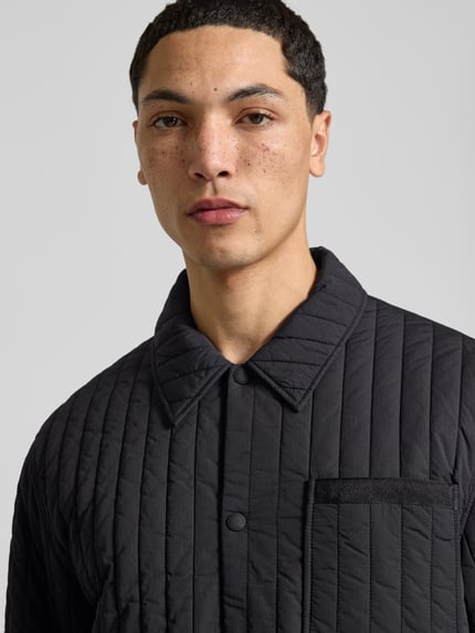 Męski model nosi kurtkę pikowaną z brustaszą marki MAISON KITSUNÉ, krój prosty, detal z logo, listwa z zatrzaskami, wpuszczane kieszenie, elastyczne zakończenia rękawów, Długość tyłu w rozmiarze M: 67 cm, Szerokość w rozmiarze M: 47 cm, Długość rękawów w rozmiarze M: 62 cm, zbliżenie na górną część torsu.