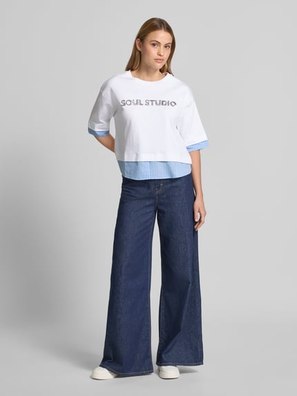 Frauenmodel trägt T-Shirt mit Paillettenbesatz und Rundhalsausschnitt von Smith and Soul aus reiner Baumwolle, Boxy-Fit, weiß mit blauen Details, Aufschrift SOUL STUDIO vorn, leicht überschnittene Schultern, Rückenlänge bei Größe S 54 cm, Ganzkörperansicht von vorne.