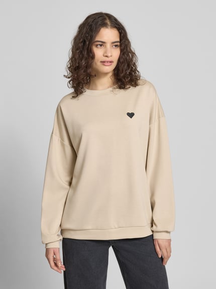 Vrouwenmodel draagt Relaxed Fit Pullover met hartborduursel model 'TALIA' van Pieces, effen beige, lange mouw, ronde hals, univormige structuur, vooraanzicht van het volledige lichaam.