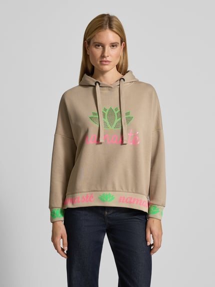 Kobiece modelka nosi Oversized Hoodie mit Strasssteinbesatz Modell 'Namaste' von miss goodlife z bawełny, krój oversized fit, kaptur z tunelem, obszycie z kamieni stras, nadruk z napisem, elastyczne wykończenia, beż i neonowe kolory, widok całej sylwetki z przodu.