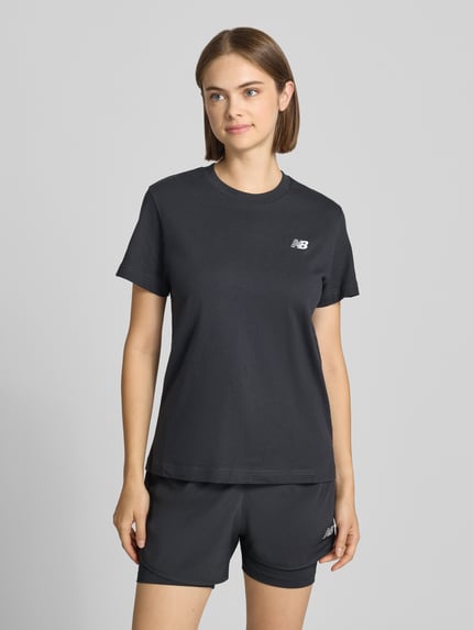 Frauenmodel trägt Regular Fit T-Shirt aus reiner Baumwolle von New Balance, schwarz, Rundhalsausschnitt, Logo-Stitching, Kurzarm, Ganzkörperansicht von vorne.