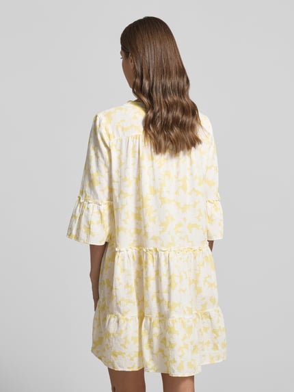 Frauenmodel trägt Ausgestelltes Blusenkleid aus reiner Baumwolle Modell 'DICTHE' von Vero Moda mit gelbem Allover-Muster, Trompetenärmel, Ganzkörperansicht Rückansicht.