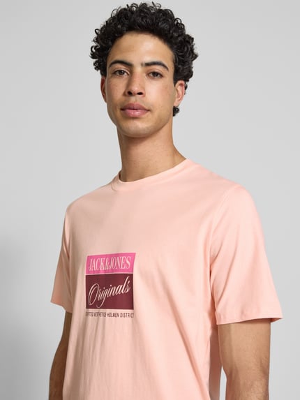 Męski model nosi jasnoróżowy t-shirt z nadrukiem z napisem model 'RHYERES' od Jack & Jones z woskowanej bawełny krój regular fit, okrągły dekolt, długość tyłu w rozmiarze M 67 cm, zbliżenie na górną część ciała