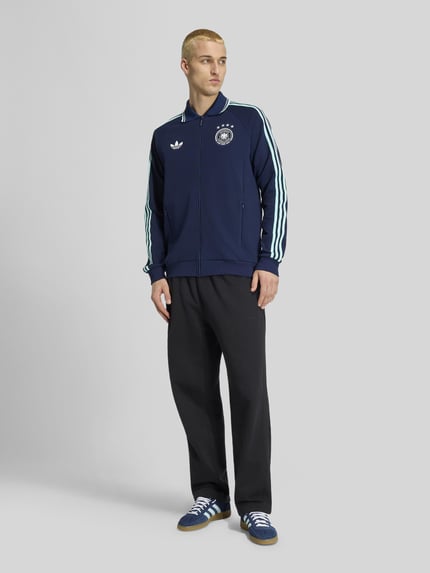 Männermodel trägt Regular Fit Trainingsjacke mit Deutschland Logo von adidas Sportswear, ciemnogranatowy kolor, kontrastowe paski, boczne kieszenie, zamek błyskawiczny, logo Niemiec z przodu, stójka, długie rękawy, mankiety ściągane, tkanina gładka; ganzkörperansicht von vorne.
