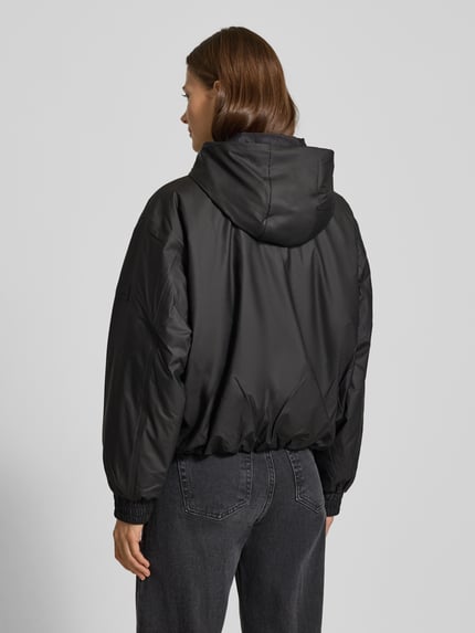 Kobiece model prezentuje czarną, luźną kurtkę z kapturem Jacke mit Kapuze Modell 'LOHJA' od RAINS, widok całej sylwetki od tyłu.