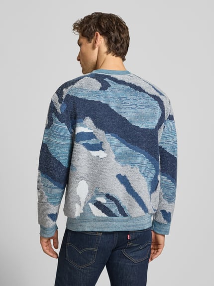 Mężczyzna nosi Loose Fit Strickpullover im Allover-Look von Scotch & Soda, bawełniany miks, niebieskie i szare wzory, krój loose fit, widok całej sylwetki z tyłu. Długość tyłu w rozmiarze M: 65 cm, długość rękawa w rozmiarze M: 60 cm.