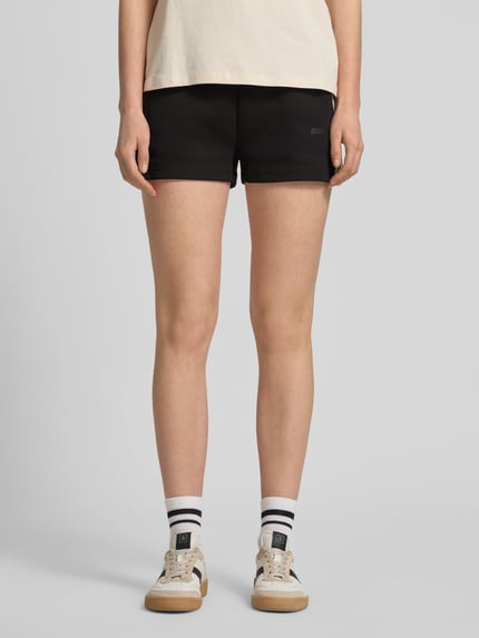 Frauenmodel trägt Shorts mit Kordelzug und Logo-Print Modell 'BRITNEY' von Guess Activewear aus schwarzem Baumwoll-Mix, Loose Fit, Ganzkörperansicht von vorne.