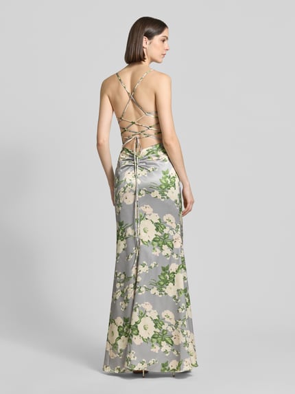Vrouwenmodel draagt avondjurk met rückseitiger Schnürung van Mascara in glanzend satijn met groen-witte bloemenprint, nauwsluitend model, maxi-jurk, cascadehals, rugdecolleté met gekruiste spaghettibandjes en drapering, ruglengte maat 36: 134 cm, borstwijdte maat 36: 33 cm. Ganzlichaamsaanzicht achteraan.