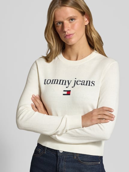 Modelka kobieca ma na sobie kremowy sweter Regular Fit Pullover aus Baumwoll-Mix von Tommy Jeans, prosty krój, miękka faktura, widoczne granatowe logo, kontekst: damski sweter od Tommy Jeans, zbliżenie na górną część ciała.