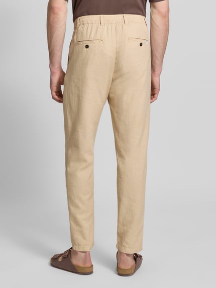 Mężczyznamodel nosi Regular Fit Hose aus Baumwoll-Leinen-Mix Modell 'BRODY' von SELECTED HOMME, jasnobeżowy kolor, tkanina melanż, etykieta, szlufki, tylne i boczne kieszenie, tunel w pasie, guziki, spodnie o kroju regular fit zwężane od kolan, zdjęcie całej sylwetki tyłem.