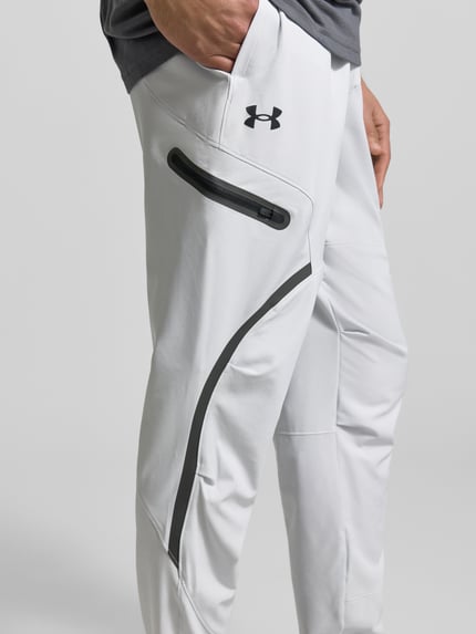 Männermodel trägt Regular Fit Sweatpants mit Logo-Print Modell 'Unstoppable' von Under Armour, elastische materiaalmix, cargozak met ritssluiting, regular fit, logoprint, elastische band, binnenbeenlengte maat M 79 cm, detailaufnahme rechter been.
