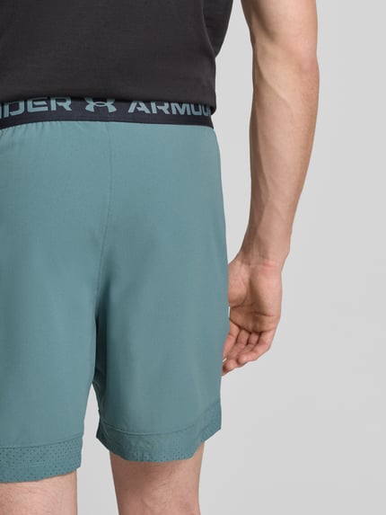 Męski model nosi Szorty z elastycznym pasem i logo od Under Armour, krój regular fit, detal z logo marki, kolor zielony, widok całej sylwetki z tyłu. Szerokość w pasie w rozmiarze M: 39 cm, długość wewnętrzna nogawki: 17 cm.