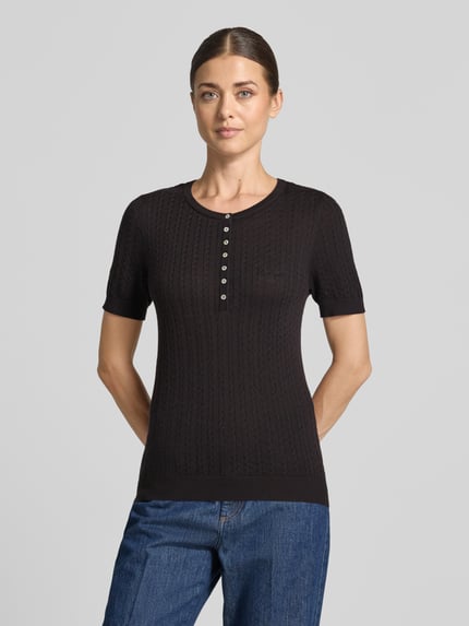Modelka kobieca nosi czarny Wollpullover aus reiner Wolle Modell 'MEADIE' od Cph Muse, dzianina z warkoczowym wzorem, okrągły dekolt z listwą guzikową, dopasowany krój slim fit, krótki rękaw z bufkami, długość tyłu w rozmiarze S: 51 cm, długość rękawa: 25 cm, widok całej sylwetki z przodu.