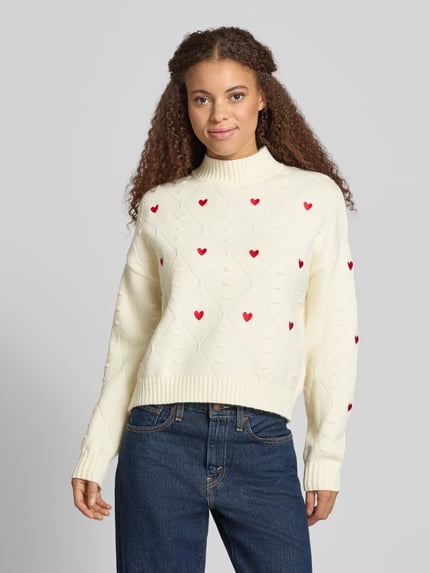 Modelka nosi Regular Fit Strickpullover mit Stitchings von NOISY MAY®, kremowy sweter z czerwonymi serduszkami, krój regular fit, stebnowania, stójka, długość pleców 52 cm, długość rękawa 53 cm, widok całej sylwetki z przodu.