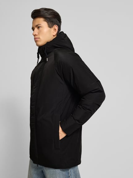 Männermodel trägt Herren Parka mit Kapuze Modell 'WOOD' von Jack & Jones in Schwarz, Regular-Fit, verdeckter Reißverschluss, Eingrifftaschen, Tunnelzug, robuste glatte Textur, Rückenlänge 84 cm, Schulterbreite 49 cm, Brustweite 61 cm, Ärmellänge 61 cm, Ganzkörperansicht von vorne.