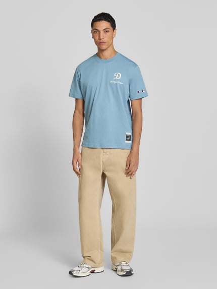Männermodel trägt Relaxed Fit T-Shirt aus reiner Baumwolle von Champion in Hellblau mit Statement-Print, ganzkörperansicht von vorne.